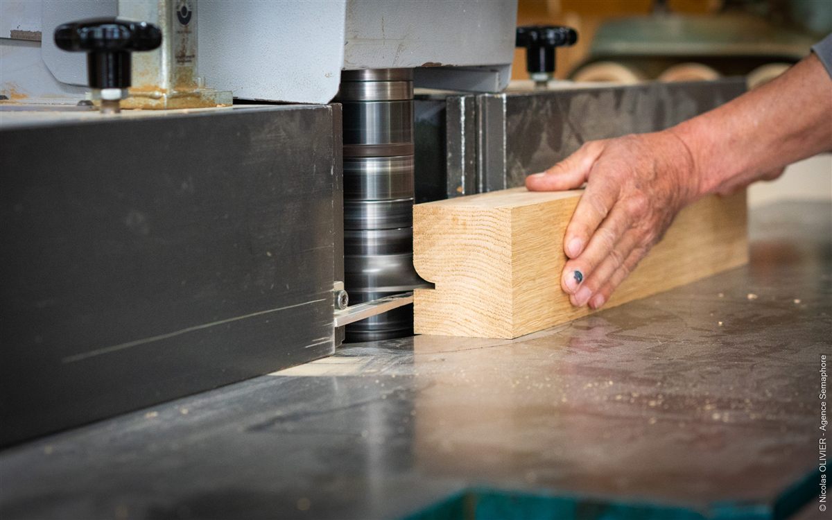 Une main guide une pièce de chêne sur une toupie, dans un atelier de menuiserie.